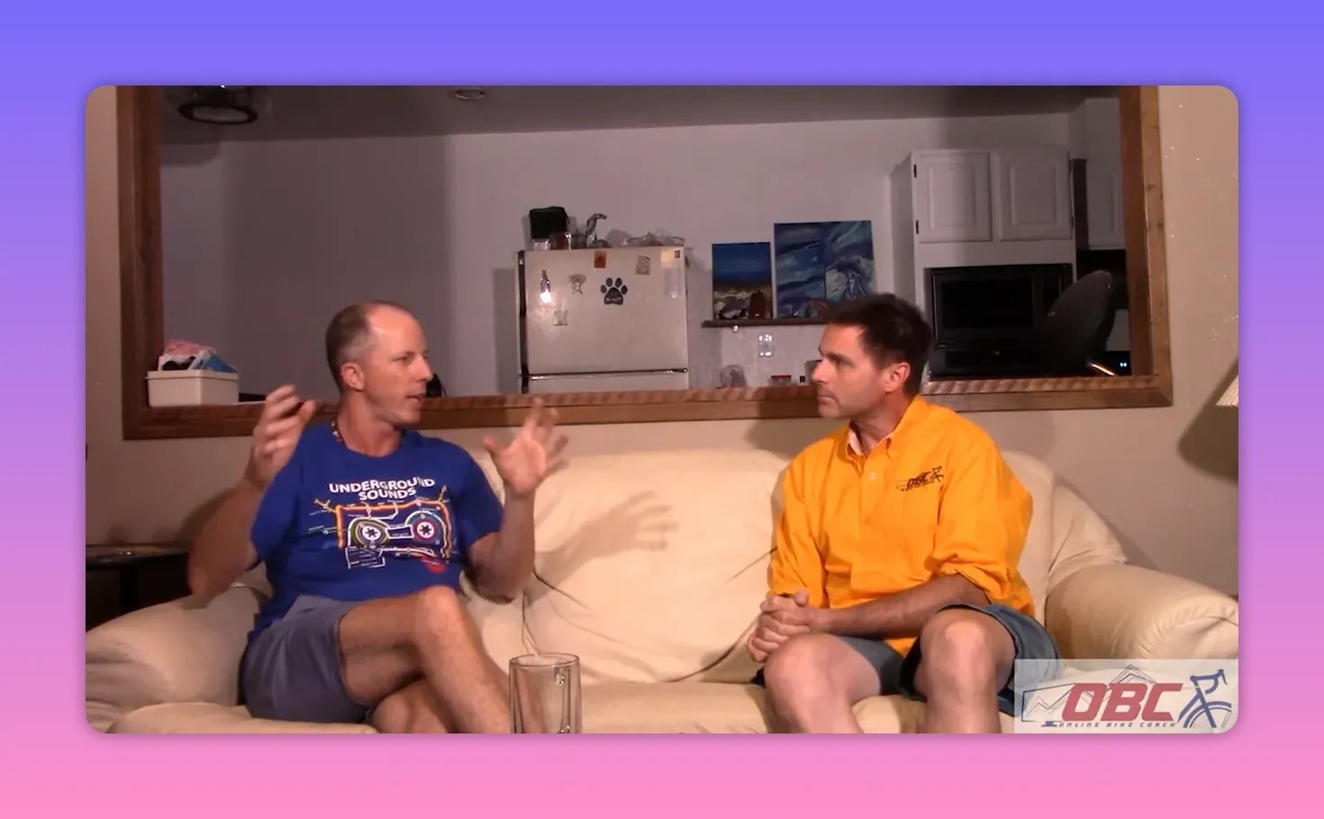 Two coaches on a couch; one uses broad hand gestures explaining training, the other listens