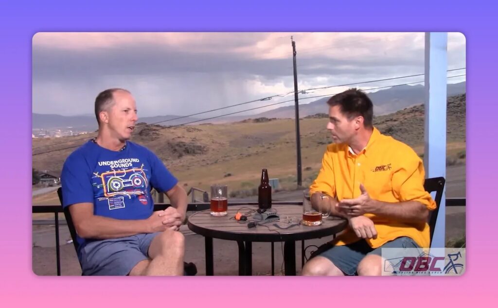 Two people in conversation at an outdoor table with rolling hills and cloudy sky behind them