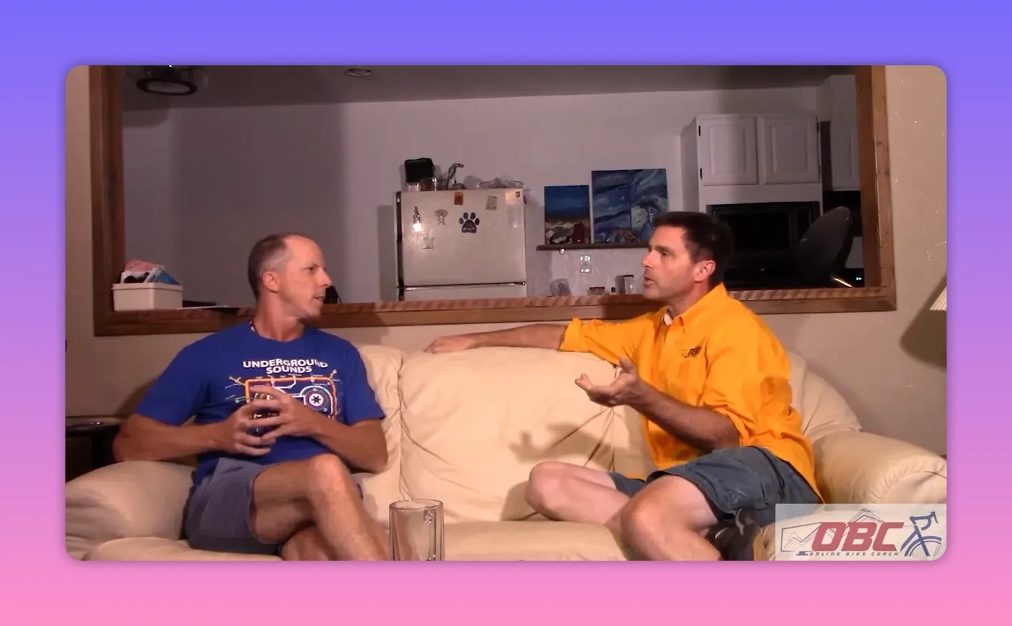 Two coaches on a couch discussing training, right-hand coach gesturing with open palm while explaining recovery concerns