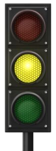 A vertical three-light traffic signal with a black housing, shown against a plain white background. The middle yellow light is brightly illuminated, while the top red and bottom green lights are dark.