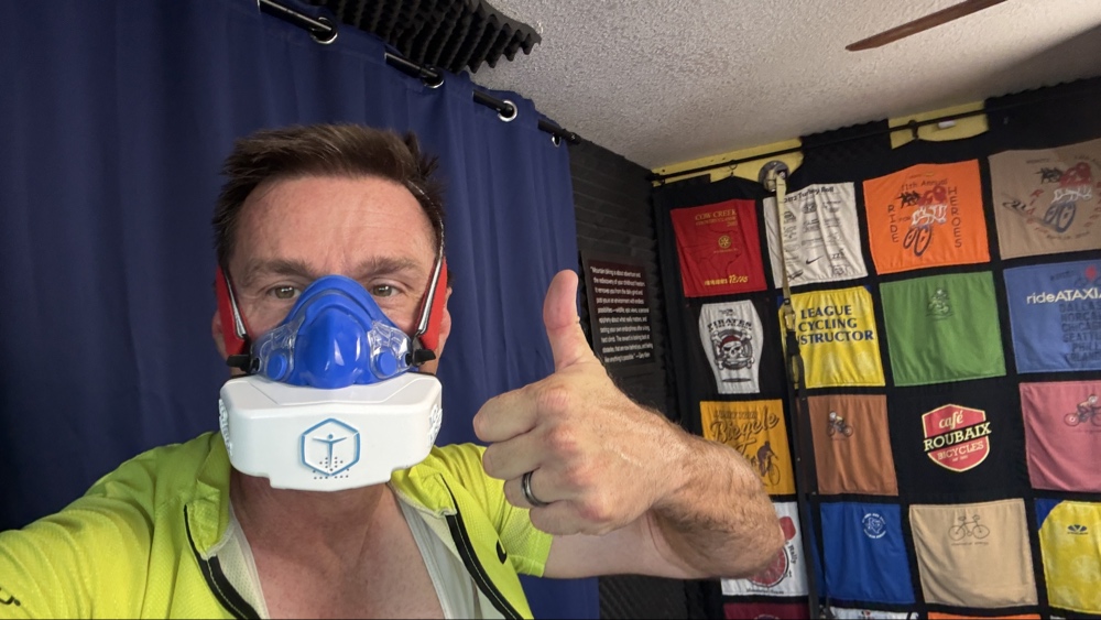 A close-up selfie of cycling coach Wharton wearing a blue and white VO2 Master metabolic testing mask while giving a thumbs-up inside his training studio, with a wall quilt of cycling event t-shirts behind him.