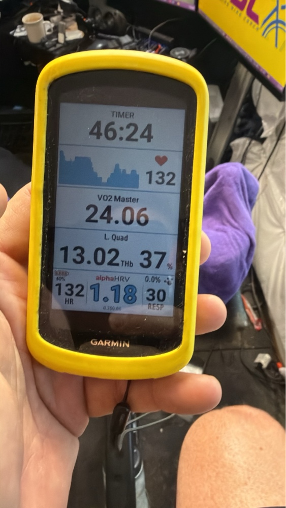 A hand holding a Garmin Edge cycling computer with a yellow protective case, displaying real-time physiological metrics including a workout timer at 46:24, heart rate at 132 bpm, VO2 Master at 24.06, left quad muscle oxygenation at 37%, and an alphaHRV score of 1.18.